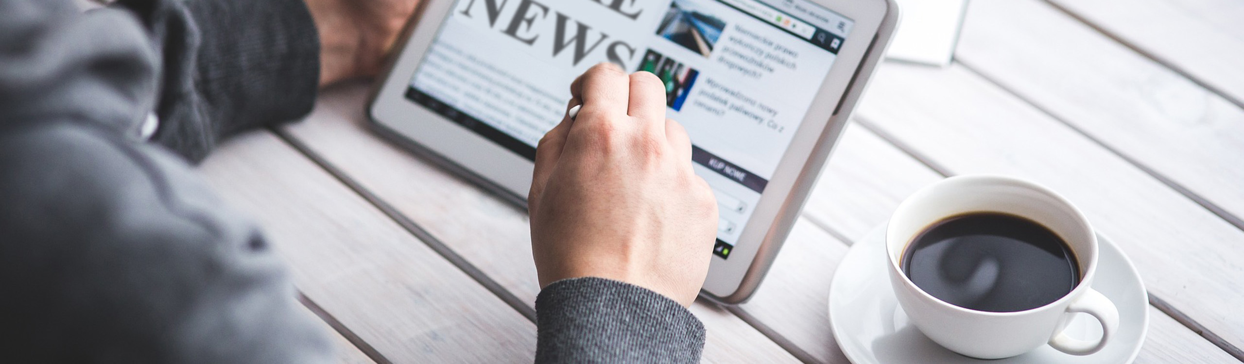 Nahaufnahmebild einer Person, die auf einem Tablet "NEWS" liest, mit einer Tasse Kaffee daneben auf einem Holztisch