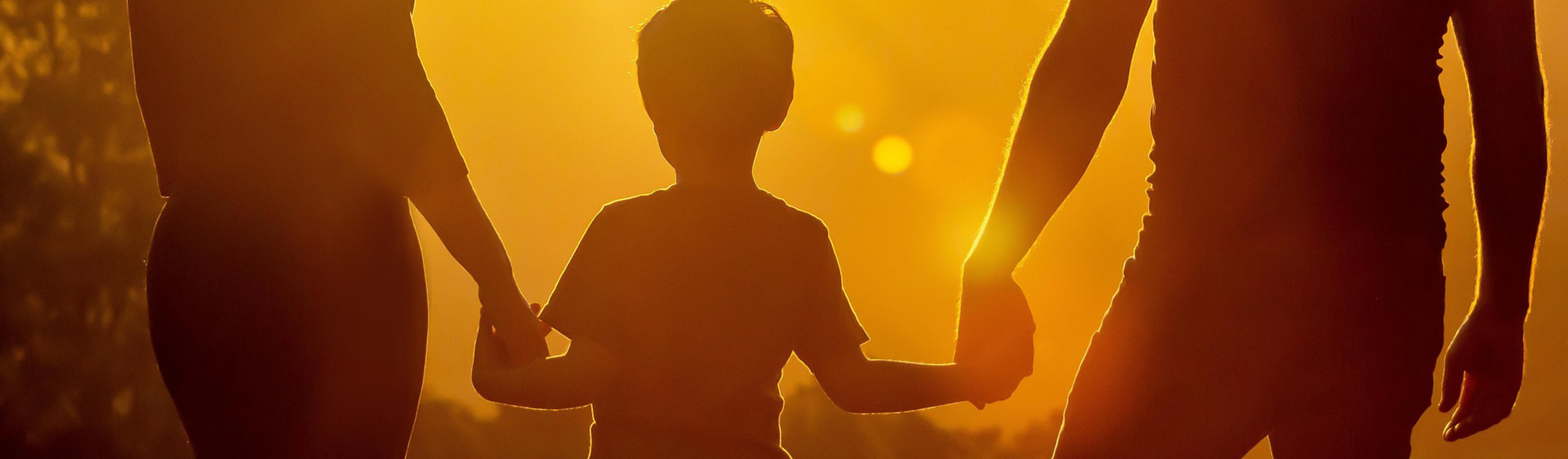 Die Silhouetten von zwei Erwachsenen und einem Kind, die Händchen halten vor einem goldenen Sonnenuntergang