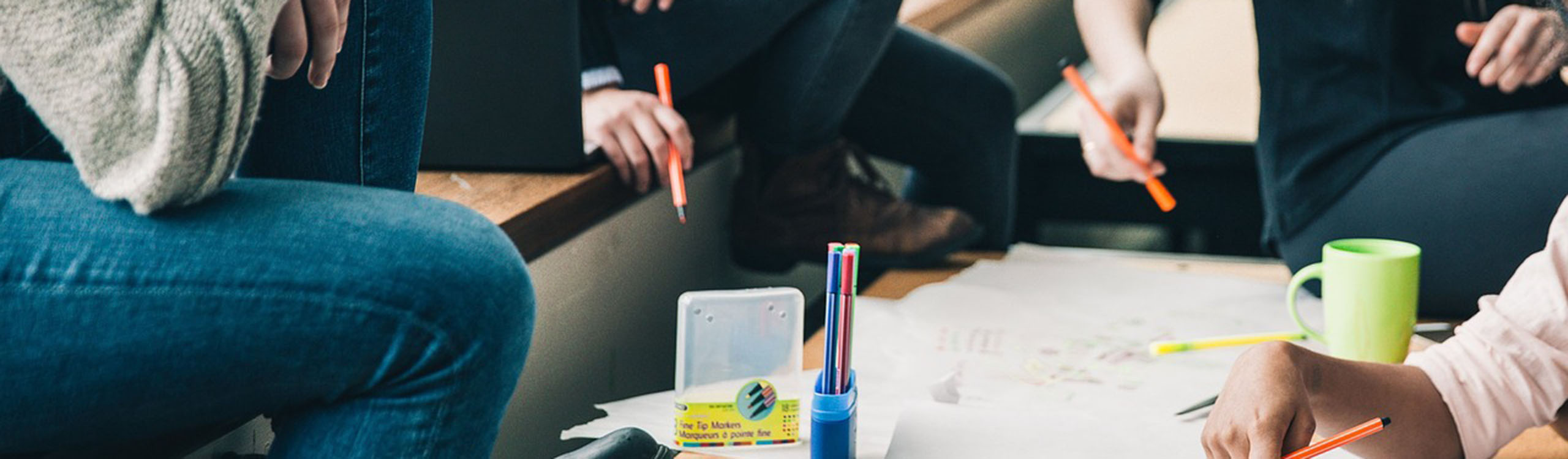 mehrere Personen sitzen um einen Tisch, auf dem Papiere und bunte Stifte liegen, und arbeiten gemeinsam