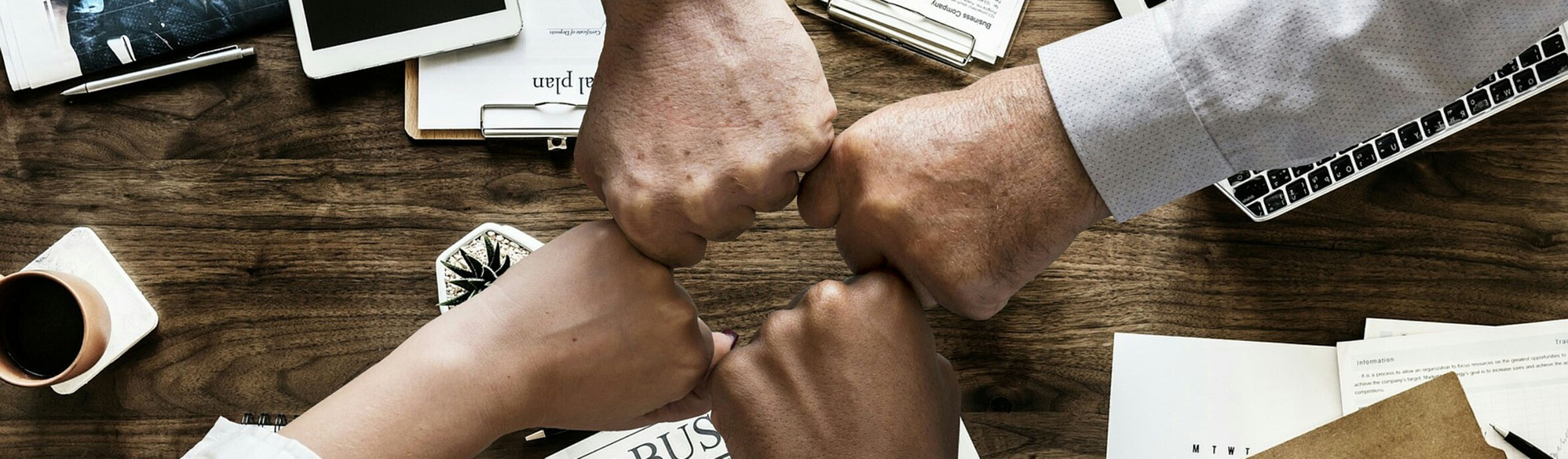 Vier Hände von verschiedenen Menschen in einem Team stoßen in der Mitte über einer Arbeitsfläche mit den Fäusten zusammen (Faust-Gruß). Im Hintergrund sind Dokumente, ein Laptop und Schreibmaterialien auf einem Holztisch zu sehen.